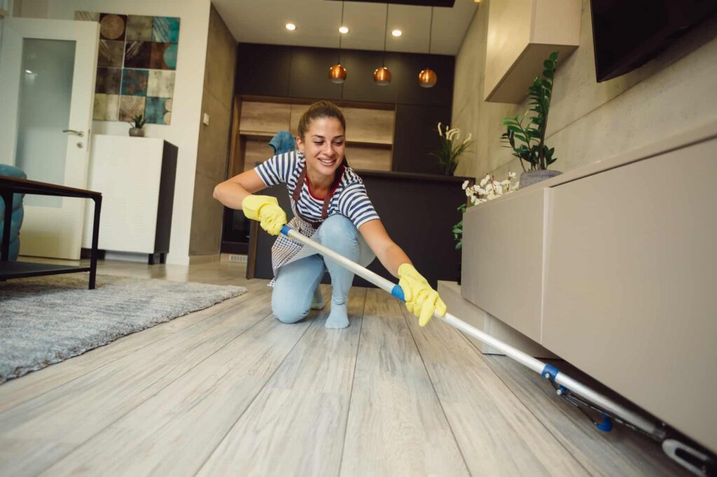 Beautiful young woman is smiling and cleaning floor under furniture at home using a mop
