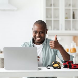 Review of construction instruments. Happy black handyman looking at laptop webcam and showing thumb